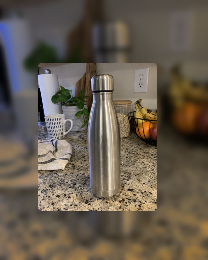 Sample photo, an AI-generated test image: a stainless steel water bottle stands on a granite countertop with a mug, paper towels, a potted plant, and a fruit bowl in the background