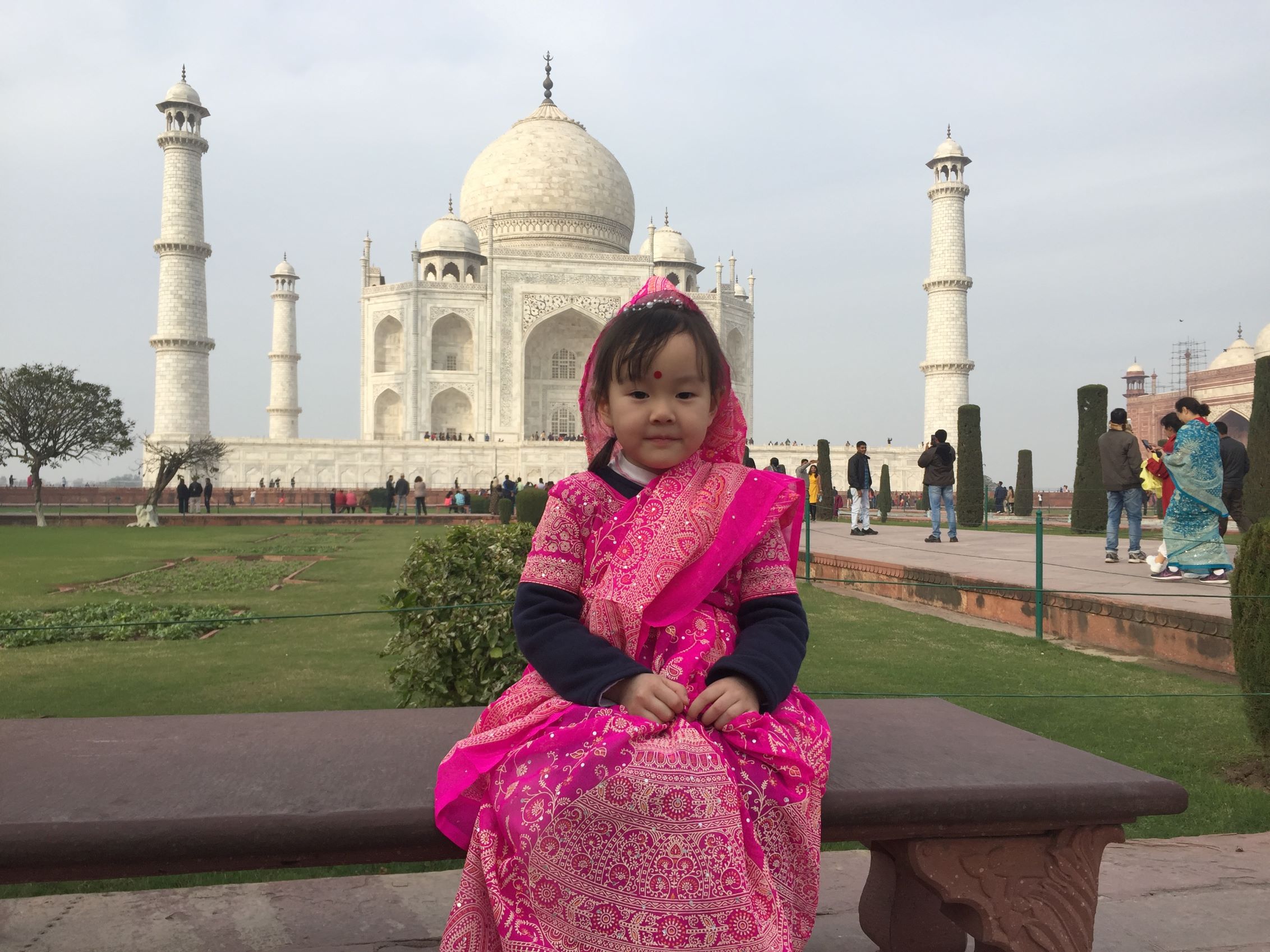 A young child sitting on a stone bench in a pink and gold traditional Indian outfit with the white marble Taj Mahal and its minarets standing in the background under a cloudy sky.