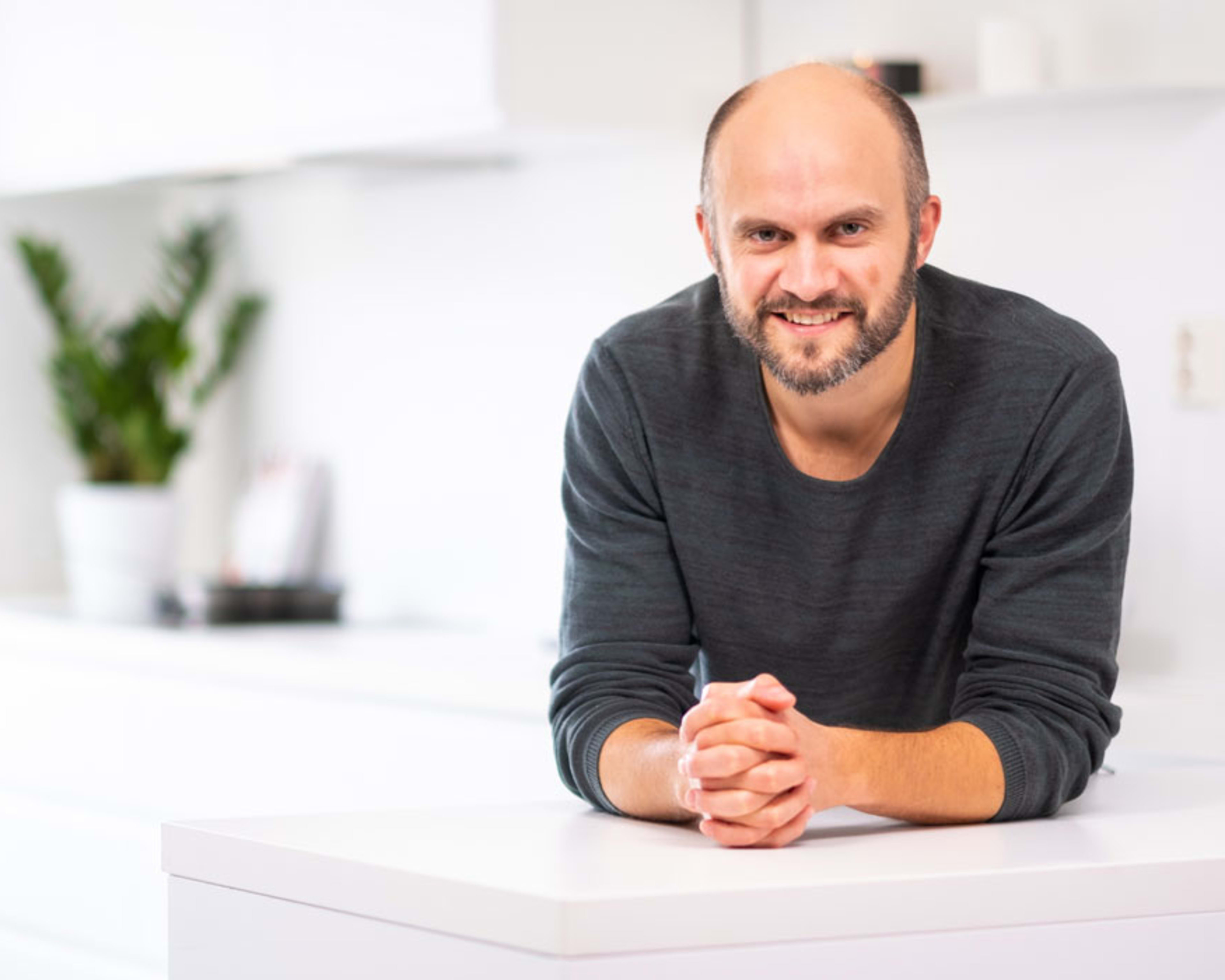 Fornøyd mann som lener seg smilende over en hvit kjøkkenbenk og ser inni kameraet.
