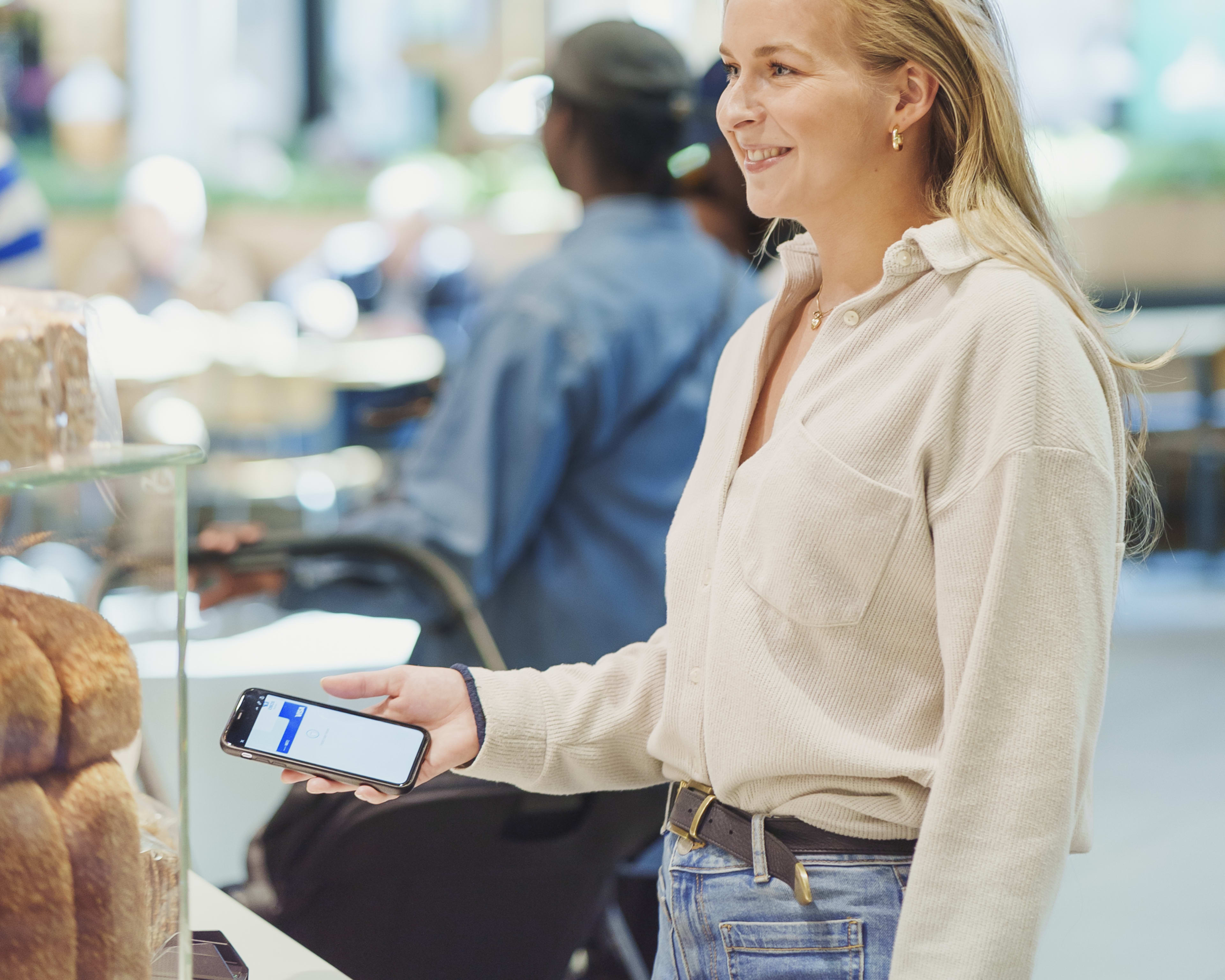 En fornøyd, blond kvinne står i kassen på et bakeri og gjør seg klar for kontaktløs mobilbetaling med Apple Pay