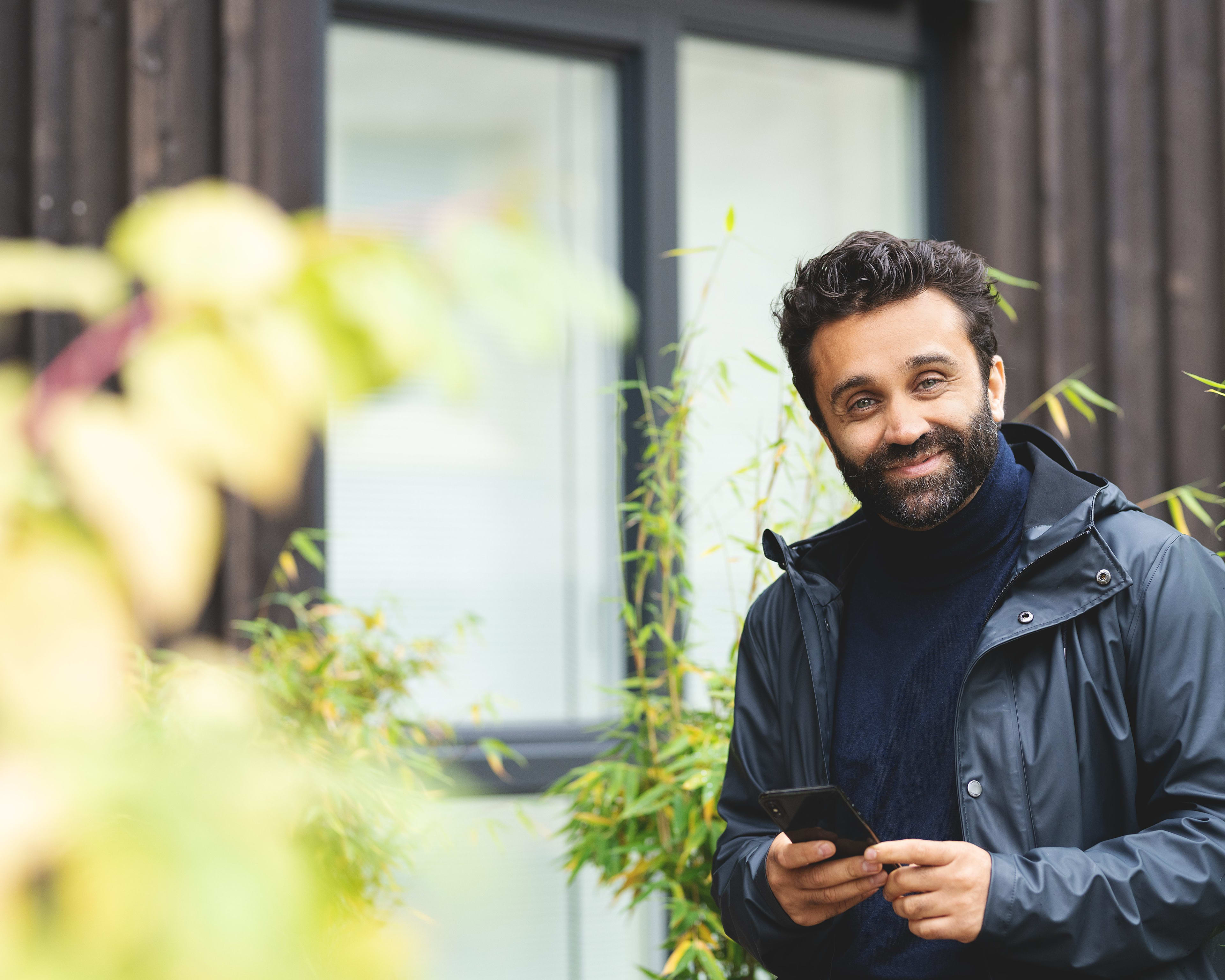 En mann ser mot kamera og smiler. Han står utenfor et hus. Det er  grønne busker rundt og han holder en mobiltelefon i hånda.