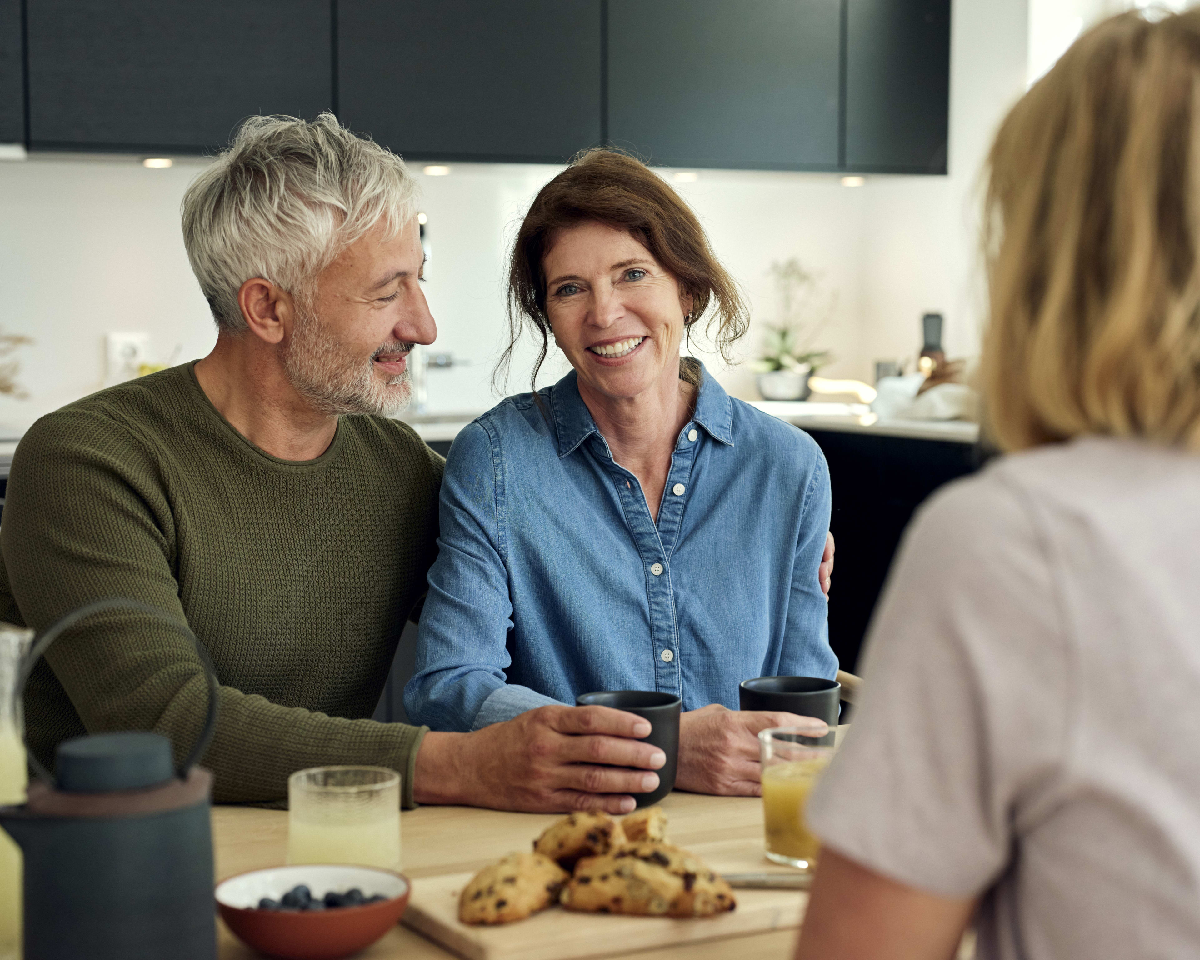 Mor, far og datter sitter rundt frokostbordet. Mor smiler mot kamera.