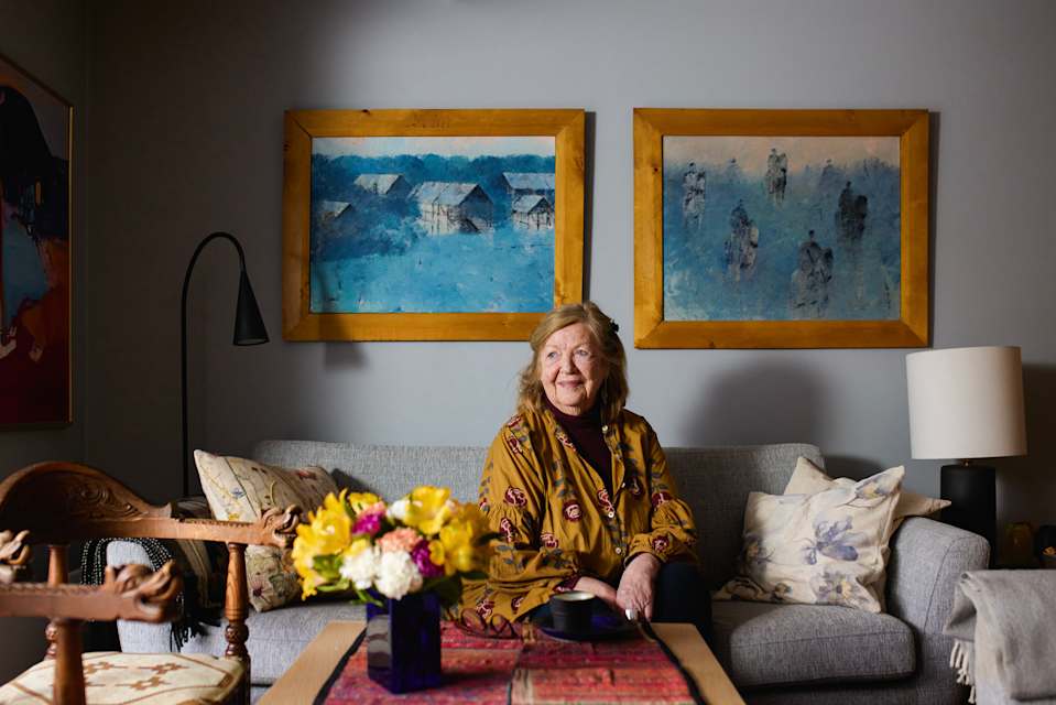 En kvinne sitter i sofaen sin hjemme. Hun titter ut og smiler. Det er to kunstverk på veggen bak henne og blomster i en vase på stuebordet.