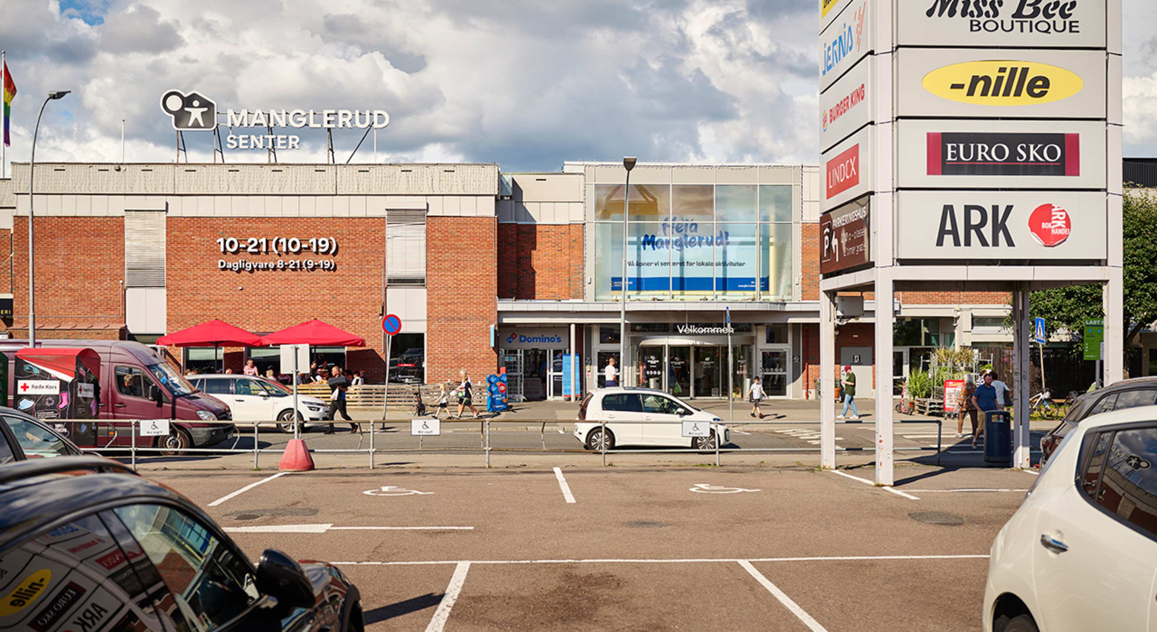 Foto av fasaden og inngangspartiet til Manglerud Senter, samt et stykke av parkeringsplassen til senteret.