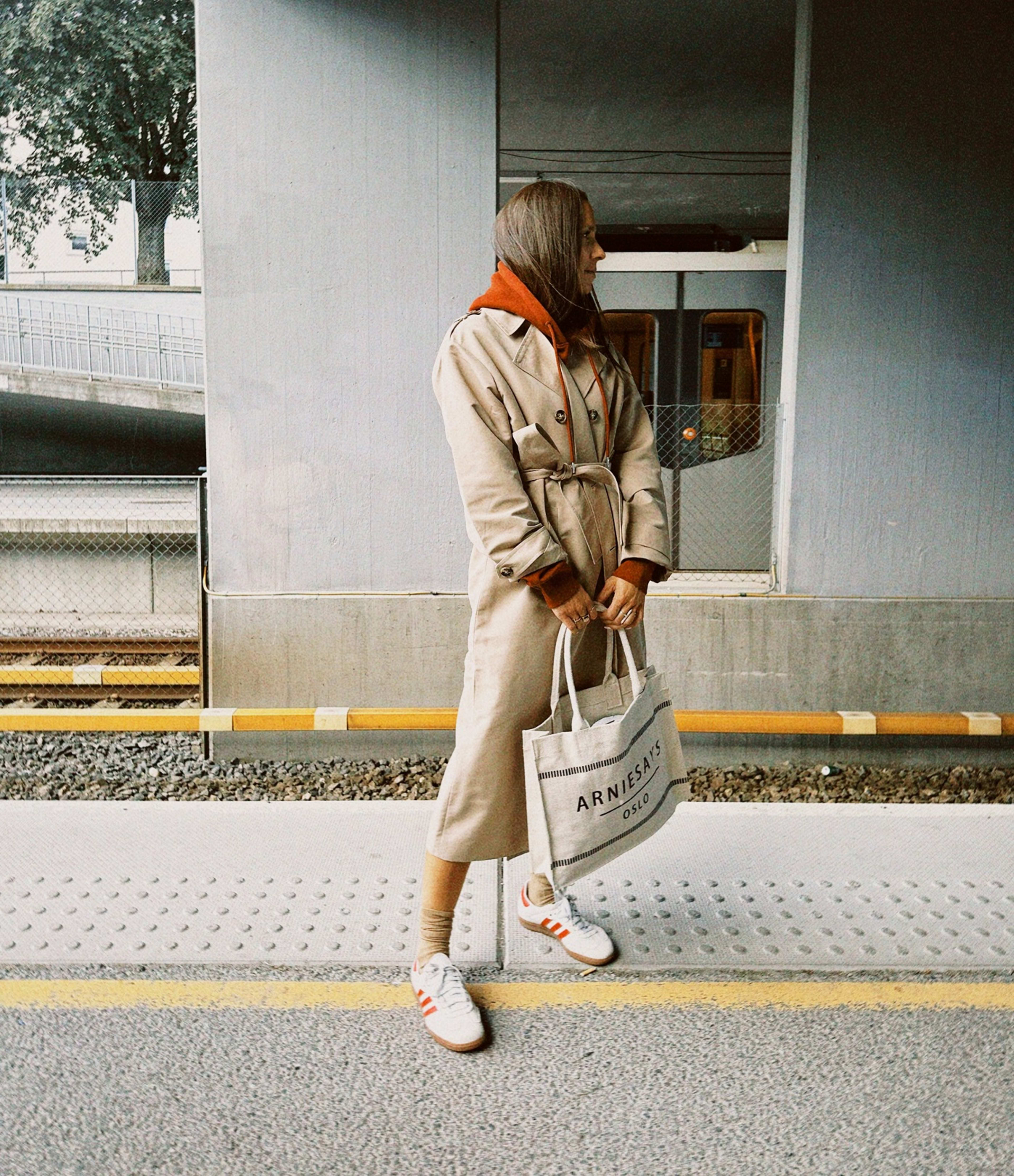 dame i lys lang trenchcoat, rustfarget hettegenser, hvite sneakers og tøyveske