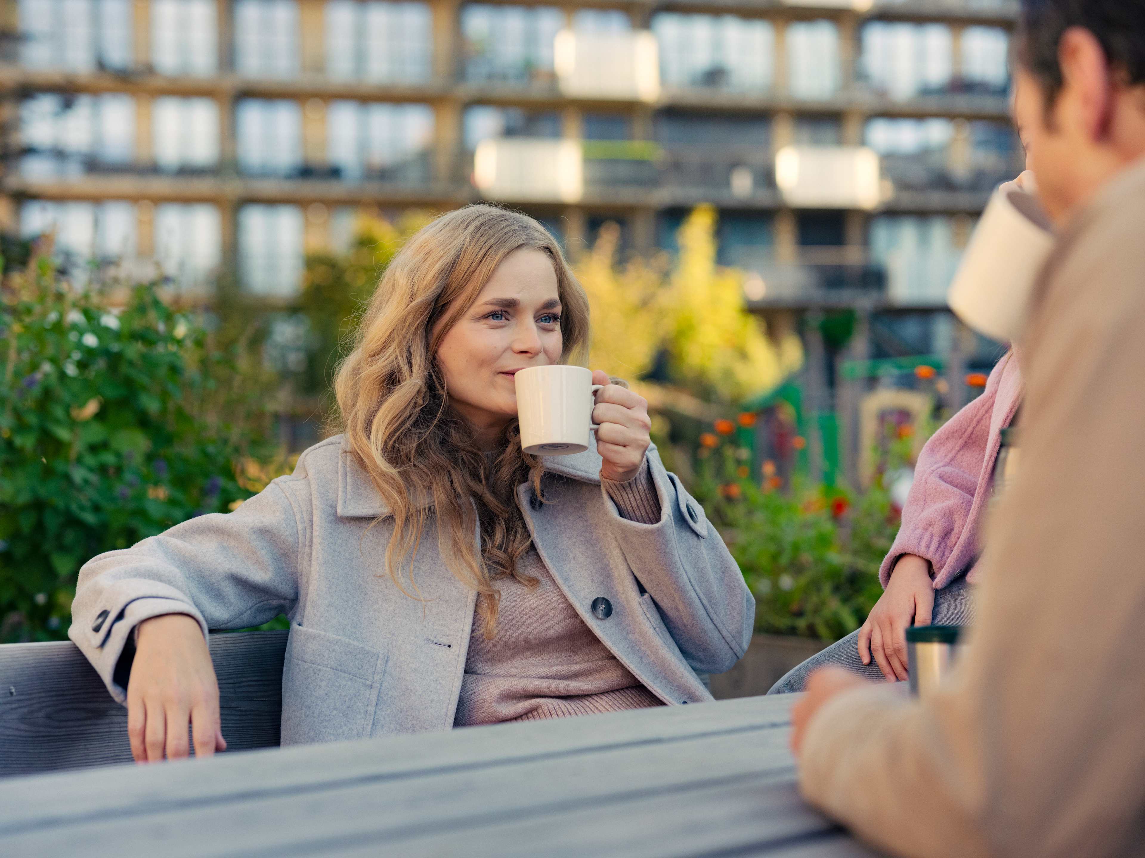 Foto av kvinne som sitter på en benk ute mellom husene, med en kopp i hånda.