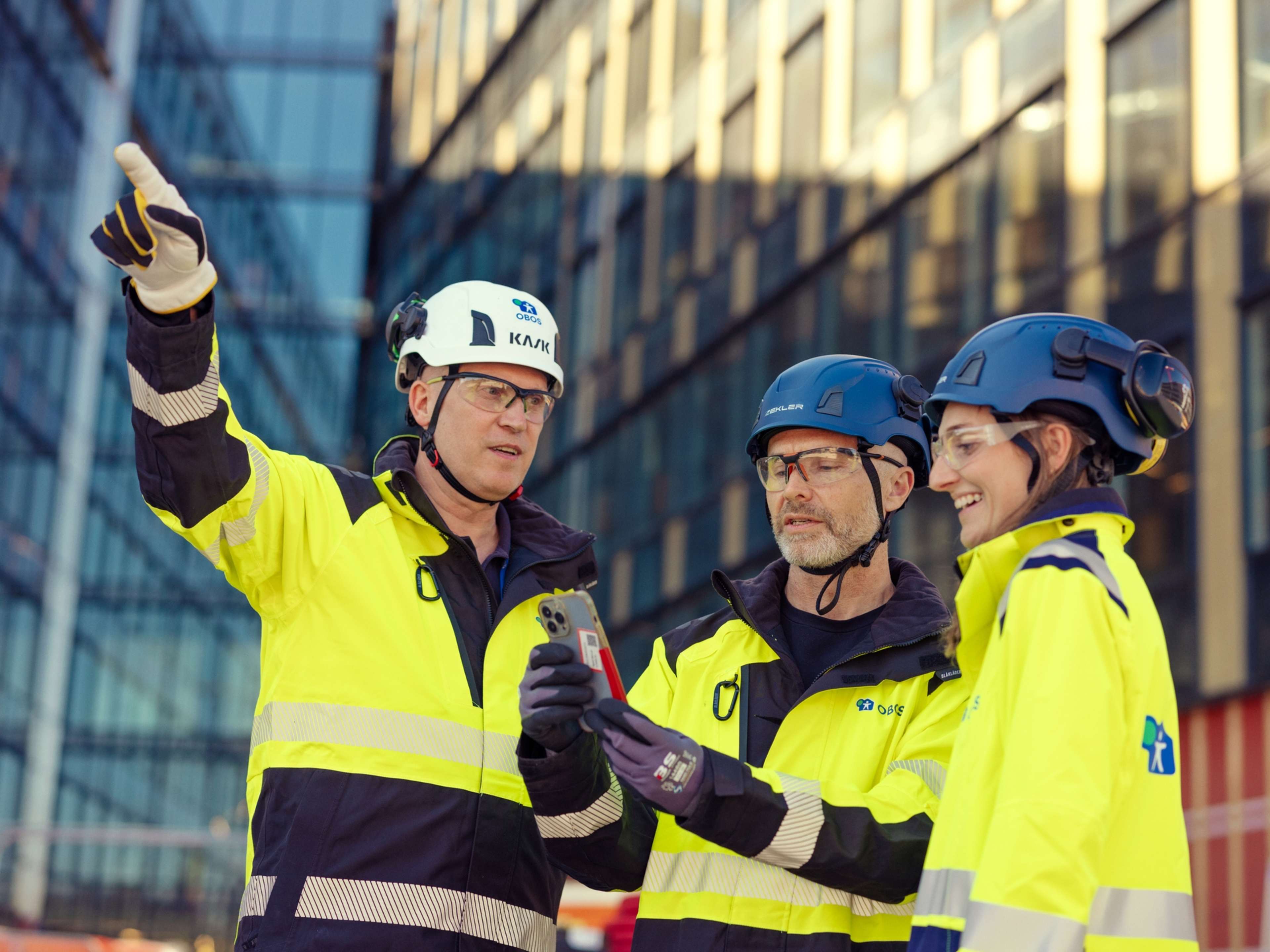 Tre arbeidere i arbeidsklær ser på en mobiltelefon utenfor et bygg under oppføring.