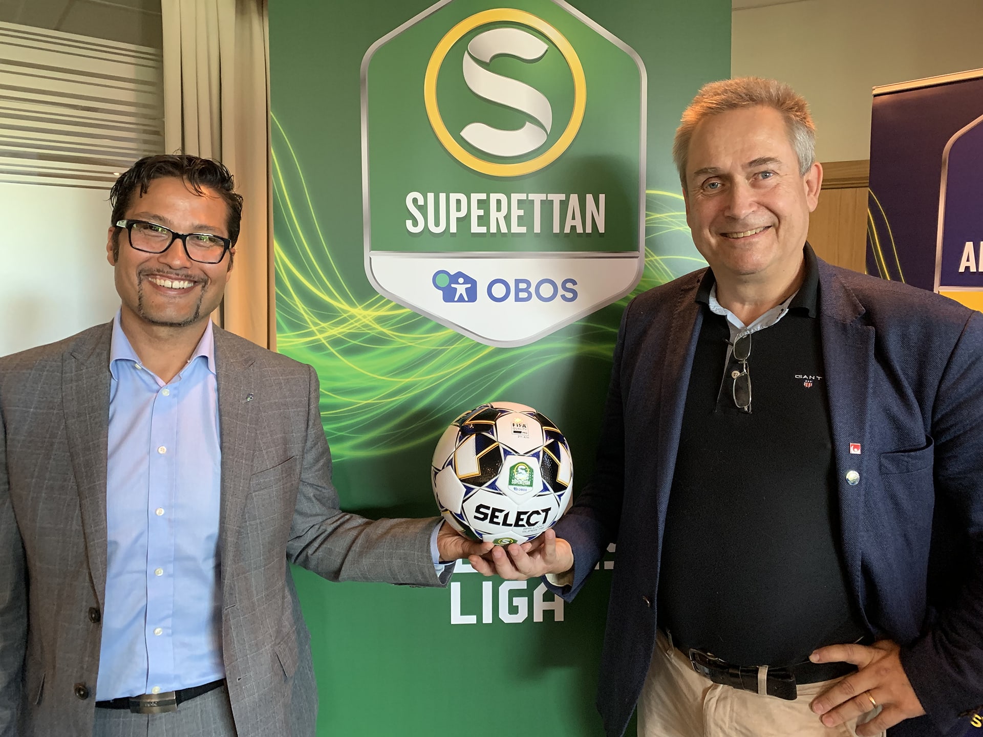 Två män står och håller en fotboll framför en superettan-vimpel.