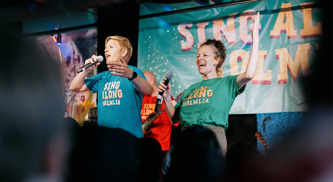 En mann og en dame fra Sing Along Holmlia står på scenen og synger foran publikum. Foto.