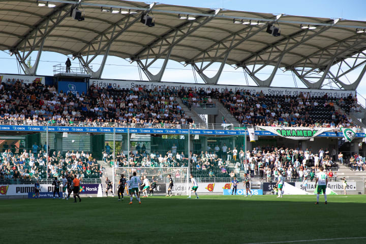 Bilde av HamKam supportere som står på tribunen rundt fotballbanen og heier på fotballspillerne på banen.