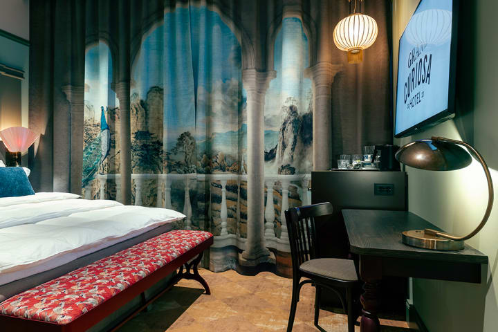 Foto av et kunsnerisk hotellrom hos Grand  Curiosa Hotel med seng, skrivebord, gardiner og en TV med hotellets logo på skjermen