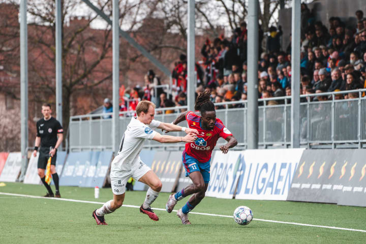 Foto av to fotballspillere som løper etter fotballen på banen med tilskuere som følger med fra tribunen