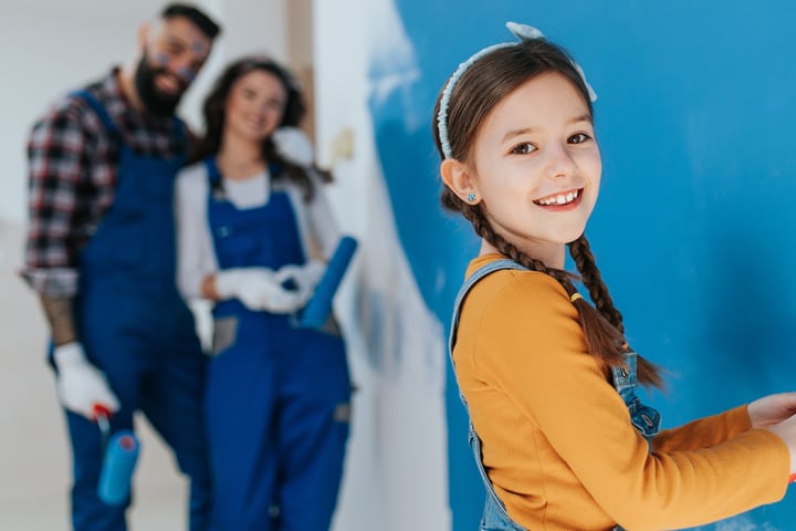 En jente maler veggen blå og smiler mot kameraet. Familien står i bakgrunnen og smiler fornøyd.