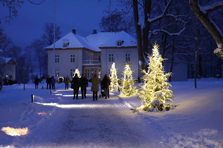 Jul i Eidsvollbygningen. Foto er tatt foran Eidsvollbygningen. Bygningen og området rundt er dekket med snø, og det går en barnefamilie mot bgningen.