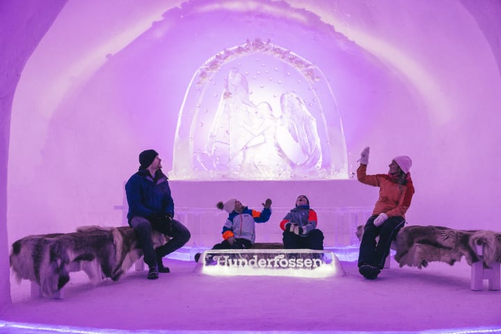 To barn og en mann og en dame sitter i en iskatedral. Det er rosa lys. Et skilt lyser opp der det står Hunderfossen. De sitter på lammeskinn for å holde varmen.