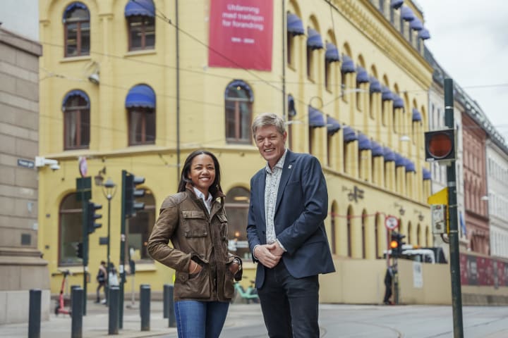 En kvinne og en mann står foran bygningen til Høyskolen Kristiania. De smiler. Bygget er gult.