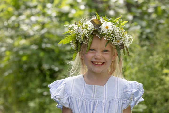 Foto av liten jente med blomsterkrans i en blomstereng som smiler til kameraet.