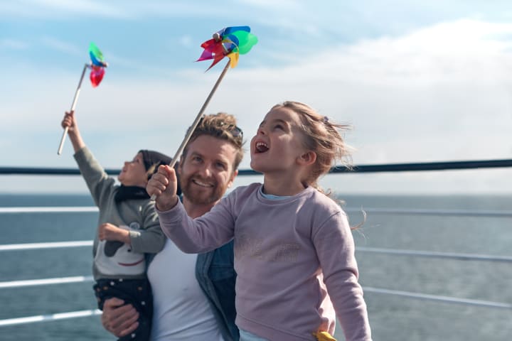 En mann og to barn smiler og gleder seg over å være om bord på dekk på DFDS. Det er fint vær og sol.