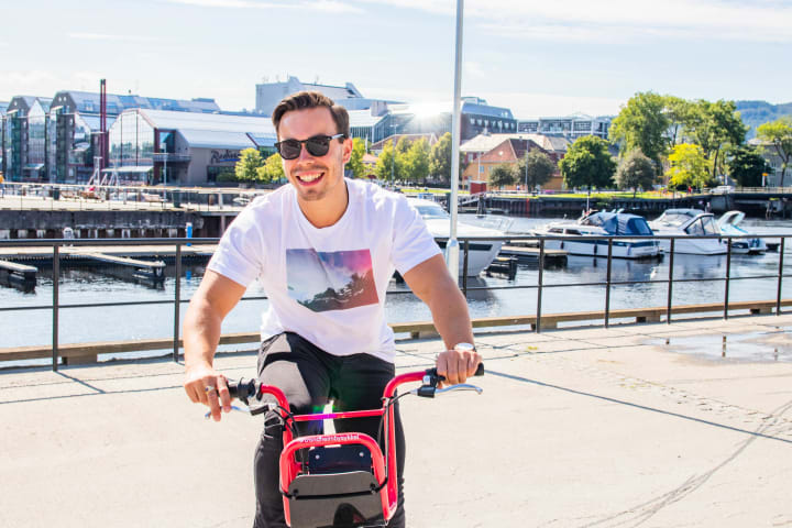 En ung mann sykler å en Trondheim Bysykkel. Det er fint vær, og han har på seg t-skjorte. Han smiler bredt og har på solbriller.