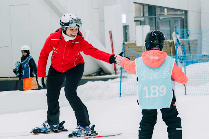 Breddebilde fra snø hvor et barn lærer å stå på slalom av en intstruktør.