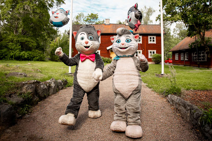 To utkledde katter vinker og sier hei. Det er et rødt hus i bakgrunnen.