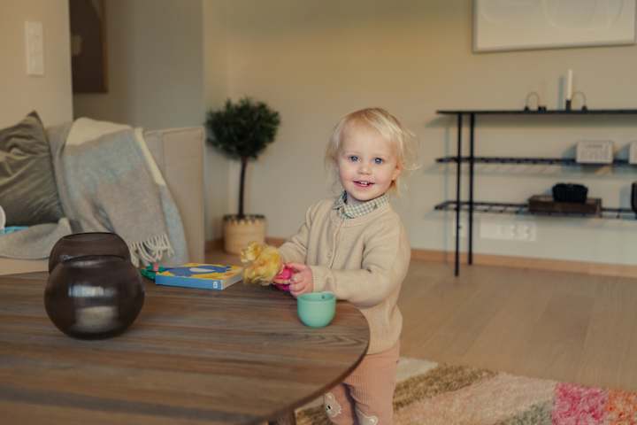 En jente har på seg beige genser og rosa bukse. Hun smiler. Hun er i en leilighet.