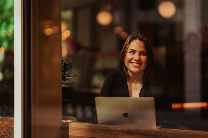 En kvinne sitter inne og ser ut av et vindu. Hun sitter med Macen. Hun smiler.