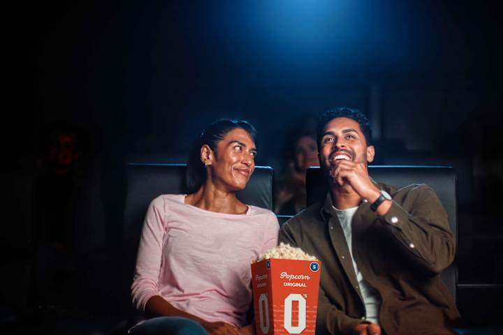 Tre barn sitter i en kinosal og ser på en film. De smiler. Hun ene jenta har popcorn og en annen jente har brus.