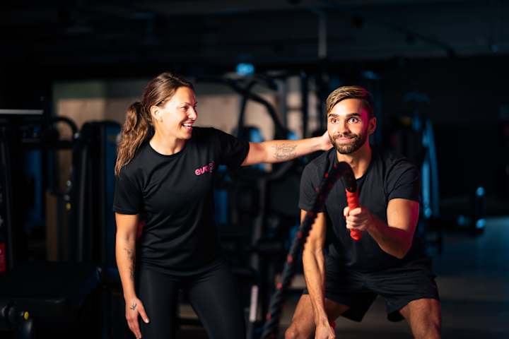 En mann trener med tau. Han holder i det og rister. En kvinnelig PT fra EVO ser på og smiler.