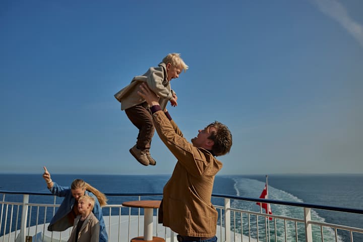 En mann løfter et barnopp i lufta. Barnet ler. Det er en familie på fire på bildet, som står på dekk på en båt.