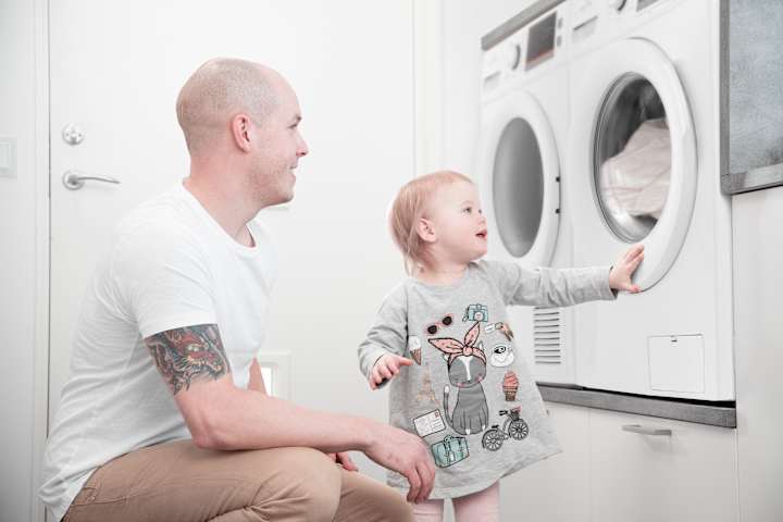 Far og barn vasker klær. Barnet ser på klærne inni vaskemaskinen.