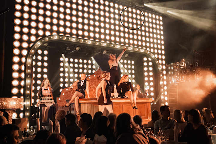 Du ser oppimot syv personer som opptrer på en scene. Damene har på seg festklær. Rundt dem sitter det folk rundt bord og ser på.