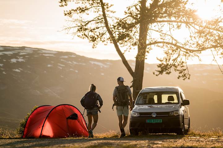 To personer ser utover fjellene. De har telt i bakgrunnen. Ved siden av teltet står en hvit varebil fra AVIS.