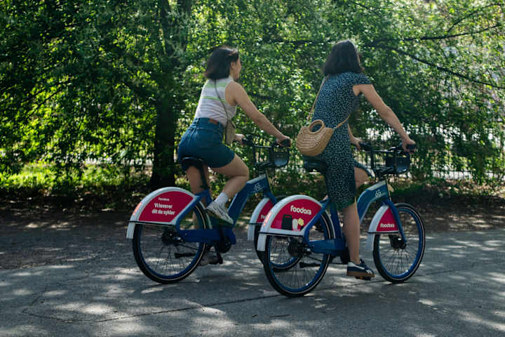 To personer sykler på en Oslo Bysykkel med Foodora på seg.