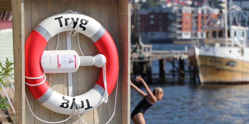 En jente hopper fra en bryggekant i vannet. I forkant henger det en Tryg livbøye