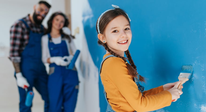 En jente maler veggen blå og smiler mot kameraet. Familien står i bakgrunnen og smiler fornøyd.