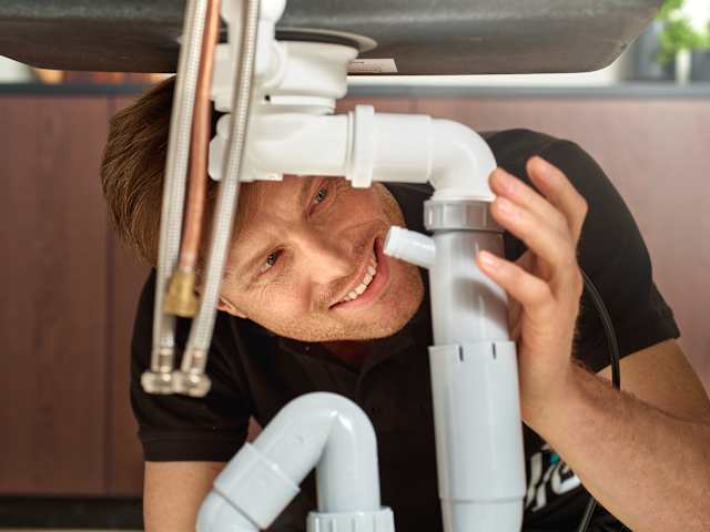 En mann står under vasken og smiler. Han ser på rørene.