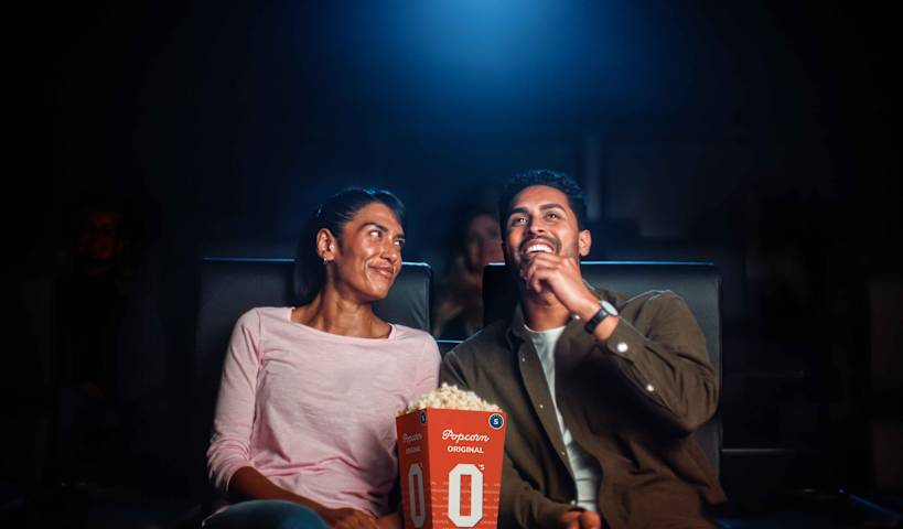 Tre barn sitter i en kinosal og ser på en film. De smiler. Hun ene jenta har popcorn og en annen jente har brus.