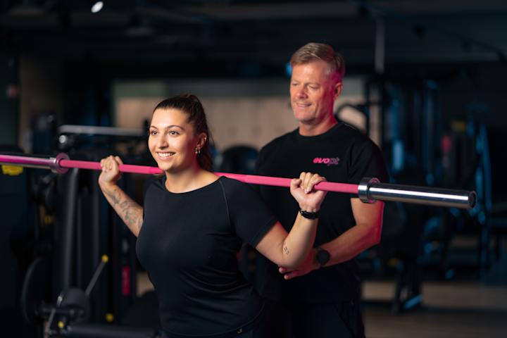 En kvinne trener med vektstang på ryggen. En mann fra EVO står ved siden og ser på. Hun smiler.
