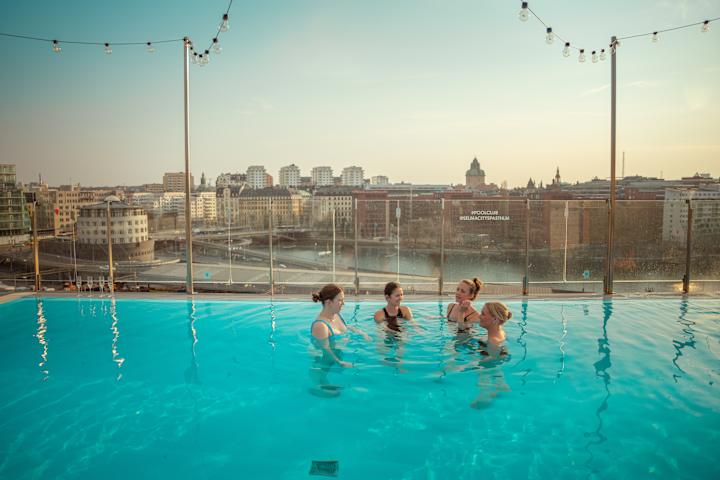 Fire damer er i et badebasseng på et hotell. De er på takterrassen. Utenfor verandaen ser du ulike bygninger. De er i en by.
