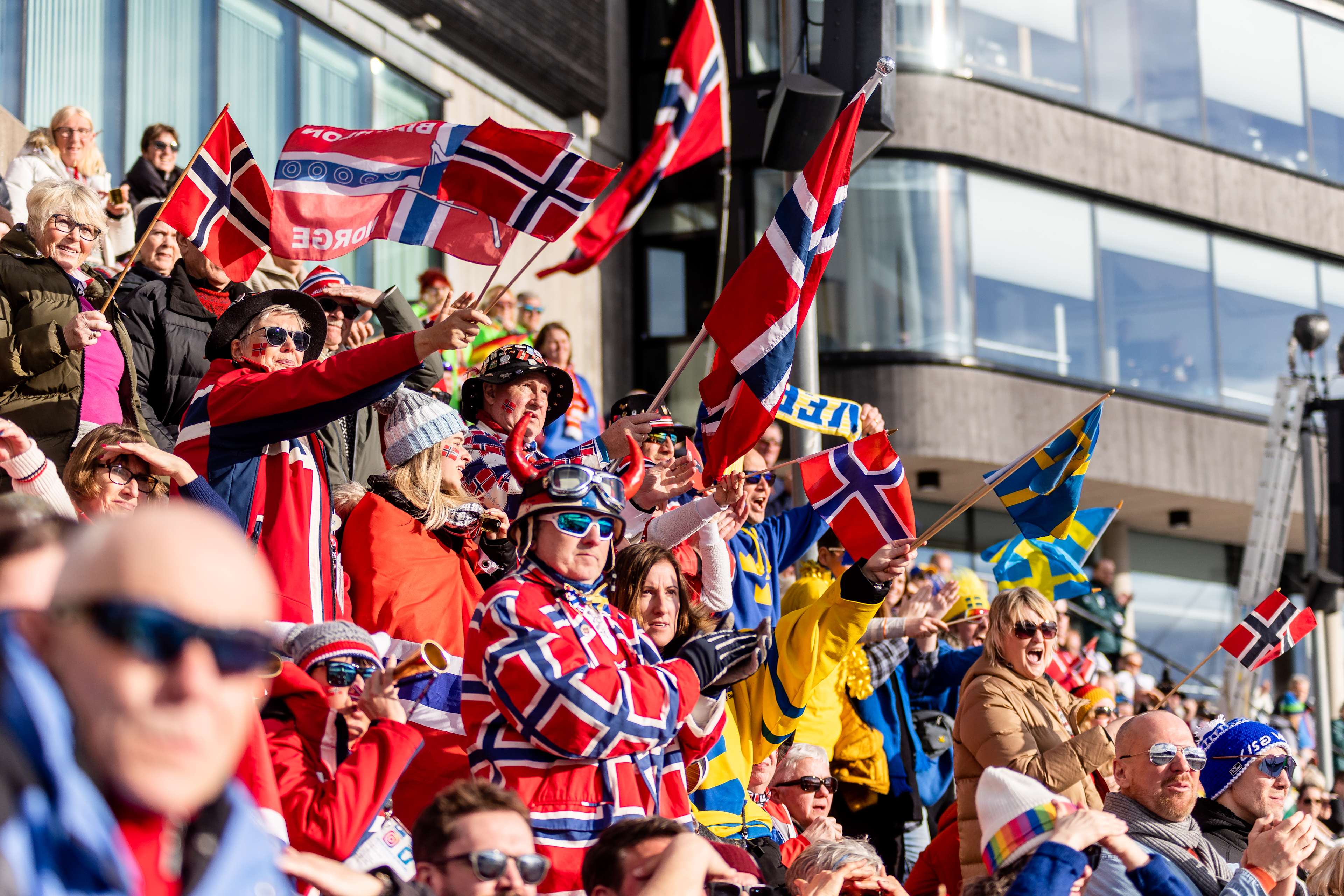 Foto av tribunen under Holmenkollen Skifest med et fargerikt publikum som heier med ulike flagg.