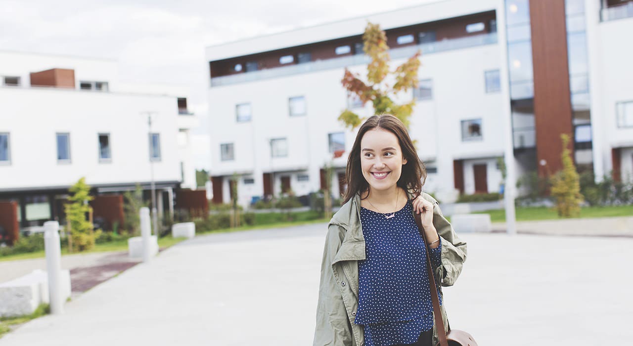 En smilende, ung kvinne er ute og går med bygninger i bakgrunnen