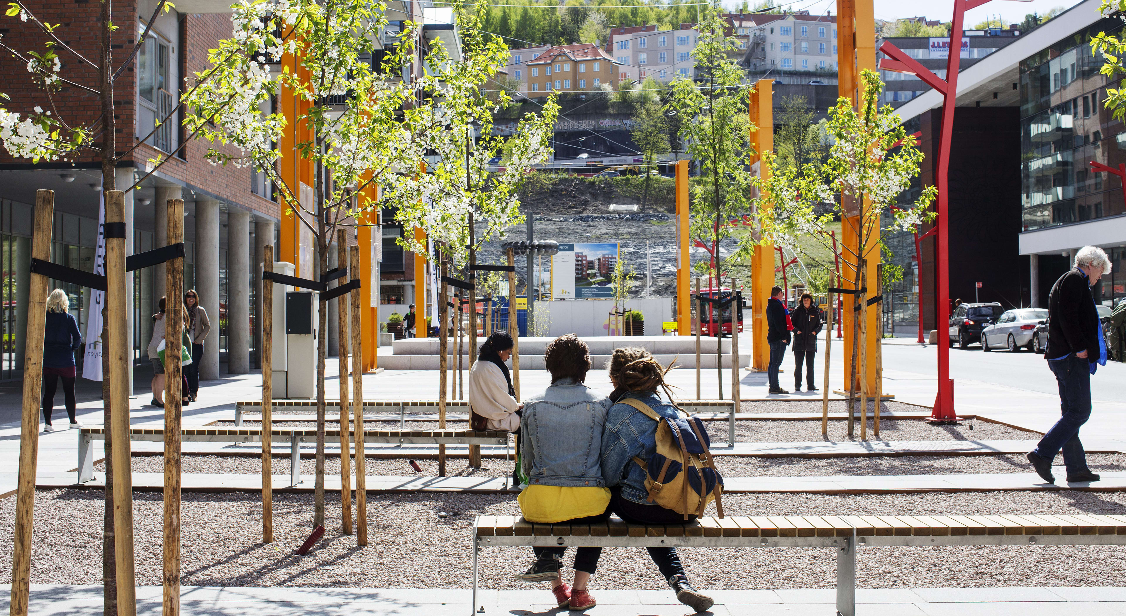 Høstbilde fra torget i Kværnerbyen. To unge jenter sitter på en benk med ryggen til, og ser ut over torget.