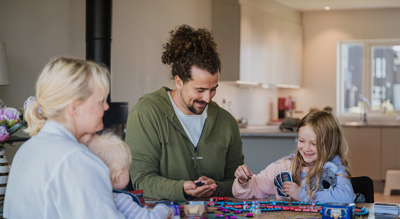 Familie på fire sitter rundt et bord og spiller brettspill. De smiler og er glade.