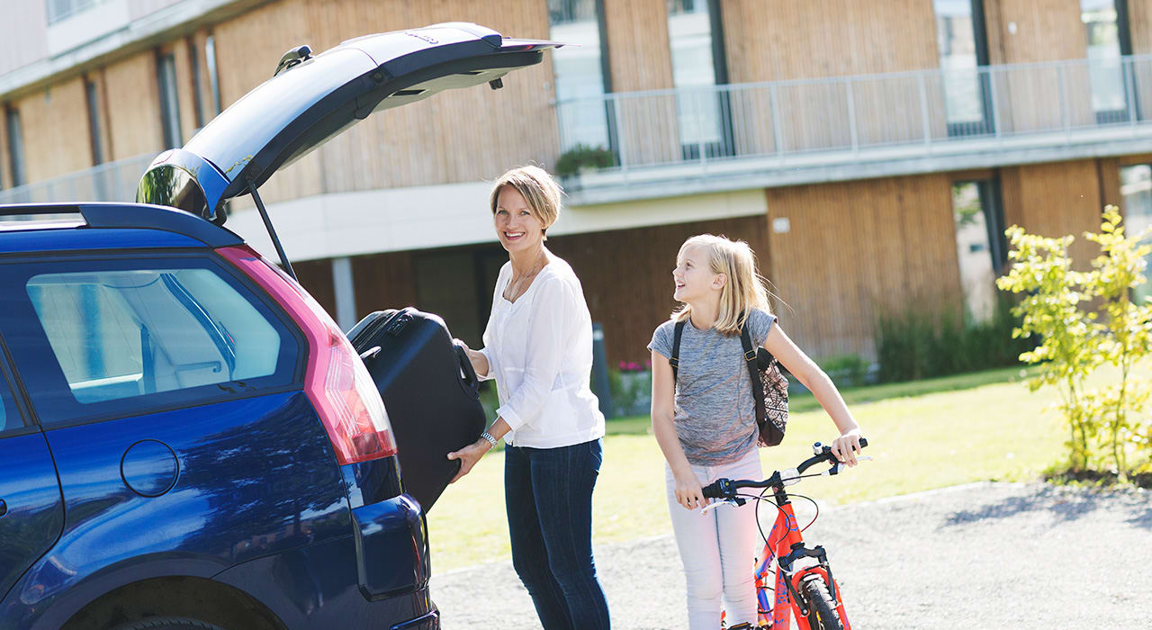 En kvinne legger en koffert i bilen og smiler. Datteren står ved siden av på sykkel.