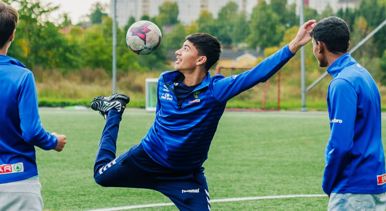 En ung gutt trikser med en fotball mens to medspillere ser på.