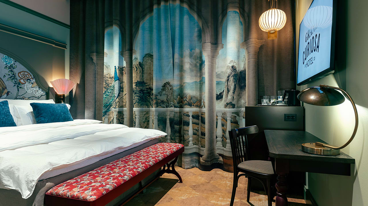 Hotellrom med en dobbeltseng, gardiner med trykk på, bord, stol og en TV-skjerm.