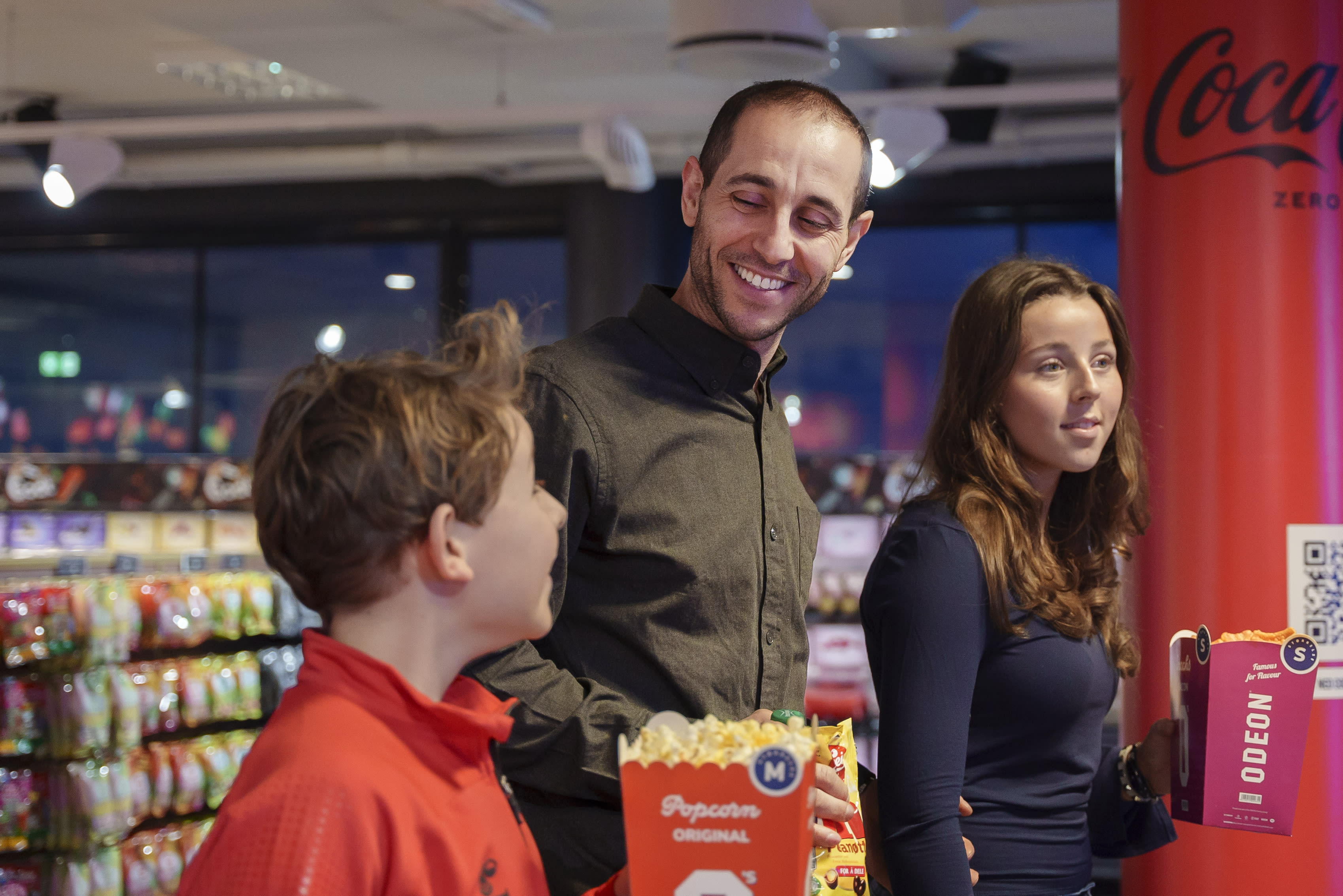 En mann skal på kino med sine to barn. De bærer hver sin popcorn. Han har på seg en grønn skjorte og smiler bredt. Barna ser forventningsfulle ut.