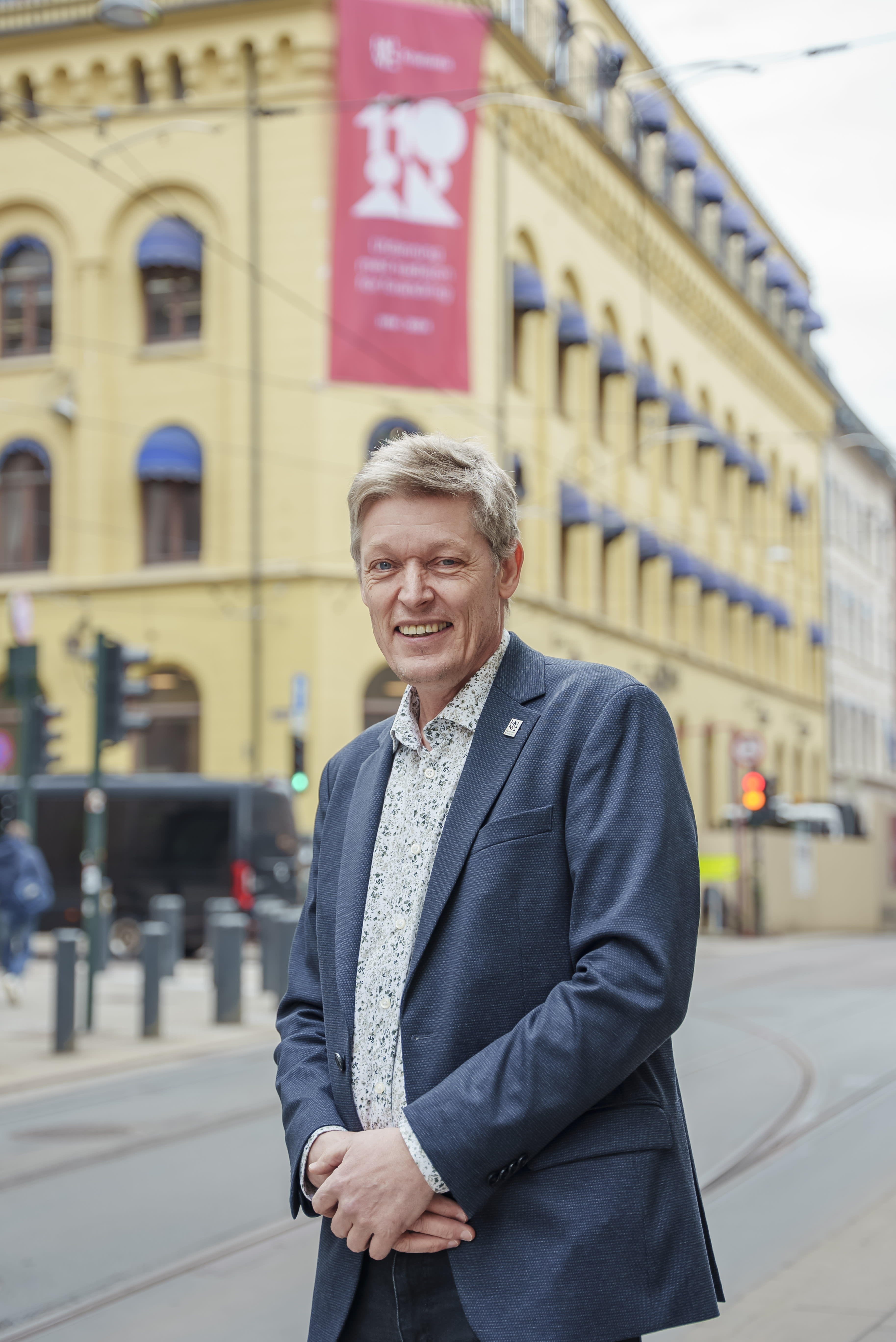 En mann står utenfor bygningen til Høyskolen Kristiania i Kirkegata. Han har på seg dressjakke. Han smiler bredt.