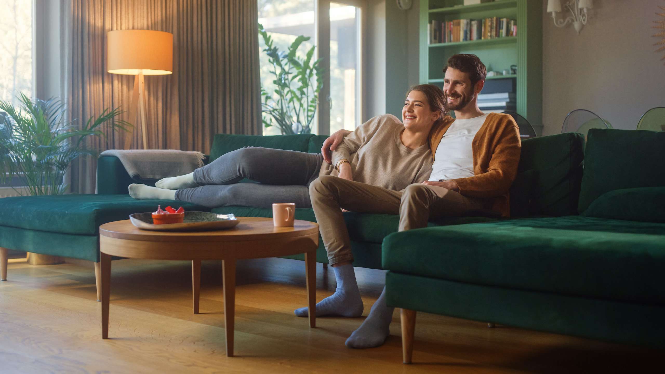 Et par sitter i en sofa og ser på TV. Hun ligger med beina i sofaen og lener seg på han. De smiler. Sofaen er grønn, og en lampe lyser.